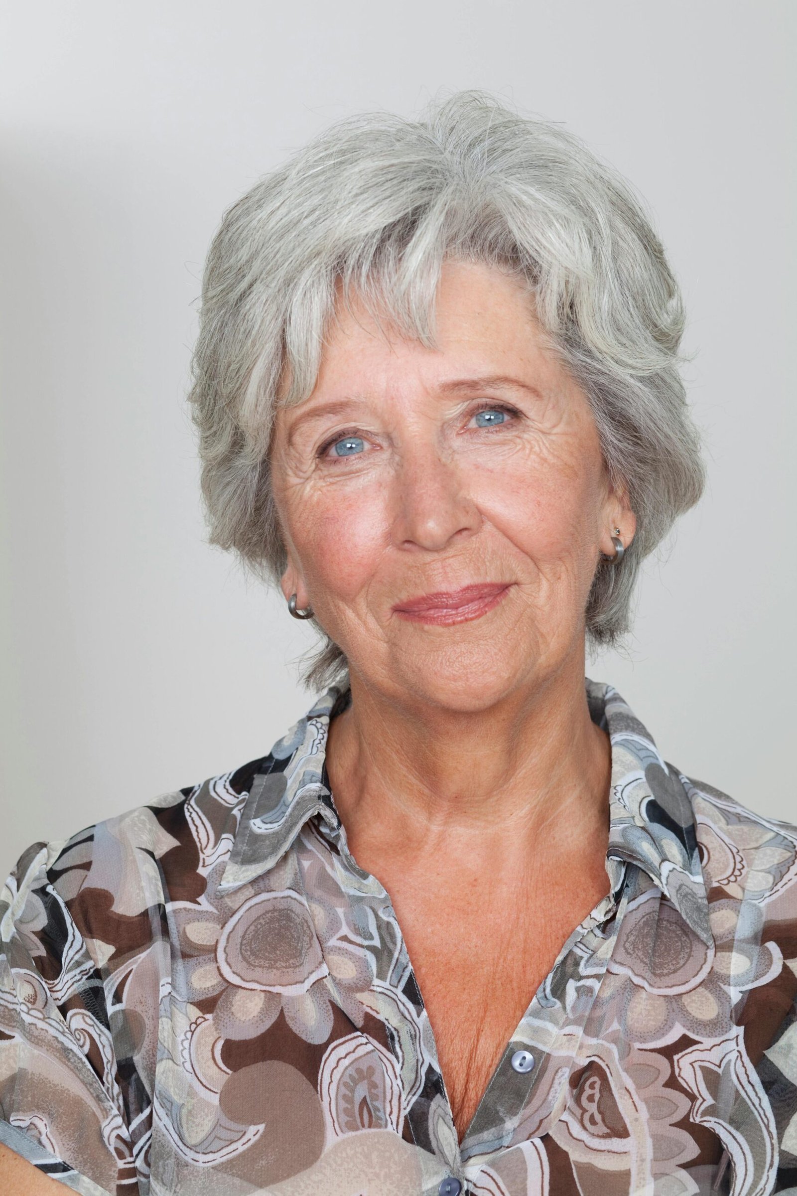 Portrait of a senior woman with grey hair in a patterned blouse on a neutral background.