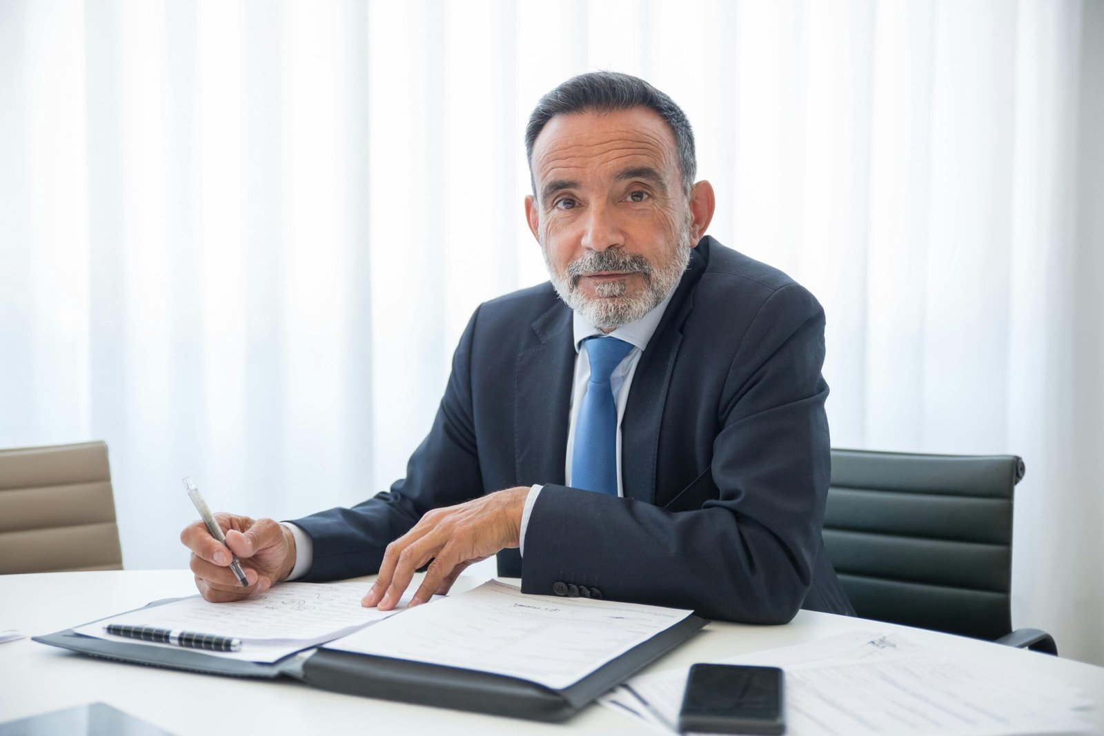 Elderly businessman with a beard signing documents in a modern office setting.