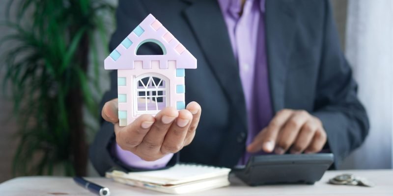 Businessman in suit holds model house, calculates real estate investments.